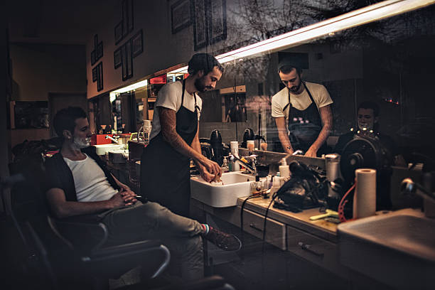 Photo of a young man, siting in a barber's chair in a little barber's shop waiting for his shaving session, while his barber is making a shaving foam in a sink
