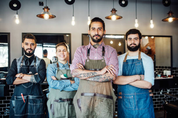 Team of hairdressers standing arms crossed in hair salon. Portrait of male entrepreneur with colleagues at barber shop. They are wearing aprons.